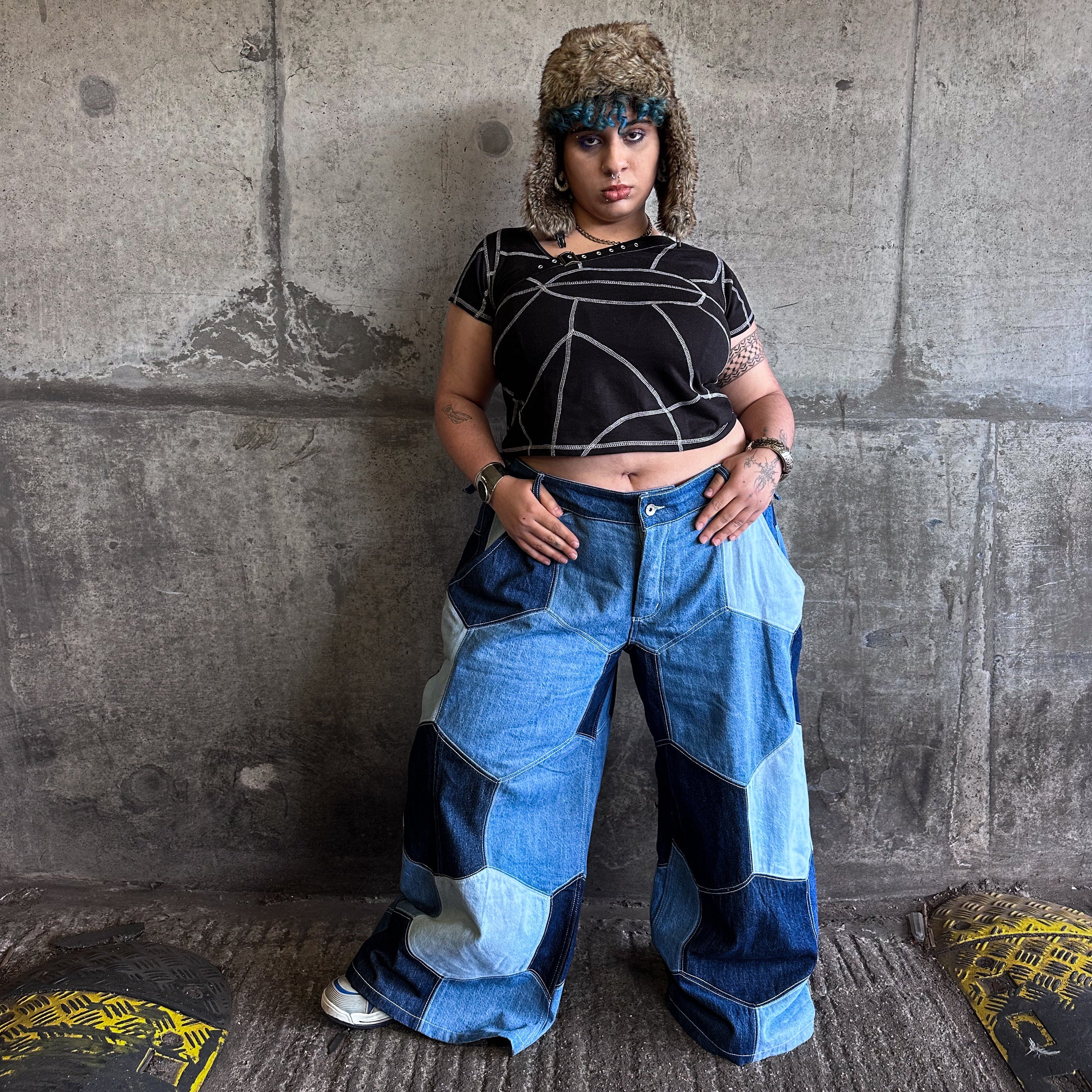 Person wearing a black buckle crop top and blue patchwork pants standing against a concrete wall.